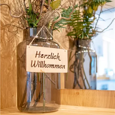 Eine dekorative Glasvase mit Pflanzen und Zweigen. Darin hängt ein Schild mit der Aufschrift "Herzlich Willkommen".