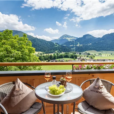 Eine gemütliche Terrasse mit Blick auf grüne Berge und eine ruhige Landschaft. Auf dem Tisch stehen zwei Gläser Rosé und frisches Obst.