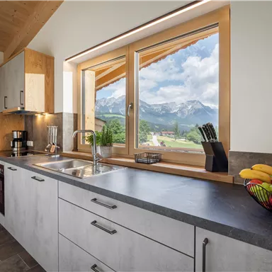 Eine moderne Küche mit einem großen Fenster, das einen Blick auf die Berge bietet. Auf der Arbeitsfläche steht eine Obstschale mit frischem Obst.