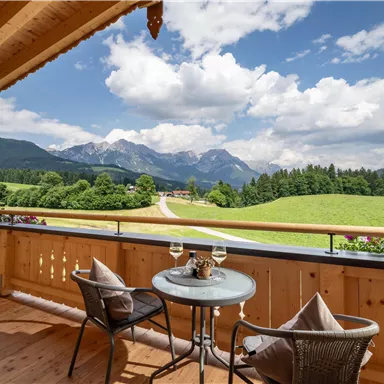 Ein schöner Ausblick von einem Balkon auf Berge und grüne Wiesen. Am Tisch stehen zwei Gläser und eine kleine Deko.