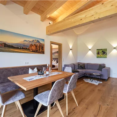 Eine gemütliche, moderne Wohnung mit Holzdecken und großen Fenstern. Der Essbereich ist mit einem Tisch und Stühlen ausgestattet, während ein einladendes Wohnzimmer mit einem Sofa den Raum ergänzt.