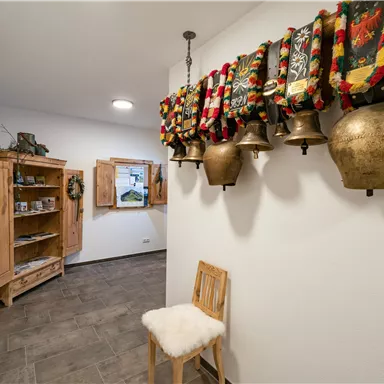 Ein heller Flur mit traditionellen Glocken und bunten Wandbehängen. Auf der linken Seite befindet sich eine Holzkommode und ein gemütlicher Hocker.