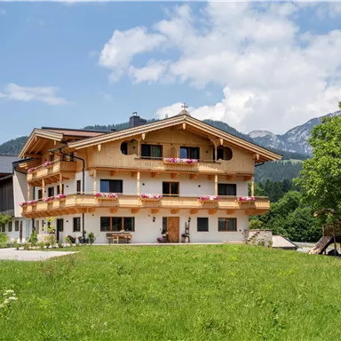 Ein schönes, modernes Alpenhaus mit Balkonen und blühendem Garten. Im Hintergrund sind Berge und ein klarer Himmel zu sehen.