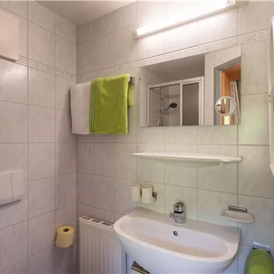 Ein modernes Badezimmer mit weißen Fliesen und hellen Handtüchern. Ein Waschbecken und ein Spiegel sind sichtbar, ergänzt durch grüne Akzente.