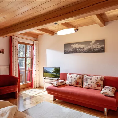 Ein gemütlicher Wohnraum mit einem roten Sofa und einem Fernseher. Große Fenster bieten viel Tageslicht und eine Verbindung zur Natur.