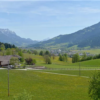 Eine malerische Landschaft mit grünen Wiesen und sanften Hügeln. Im Hintergrund sind die Berge und ein kleiner Ort zu sehen.