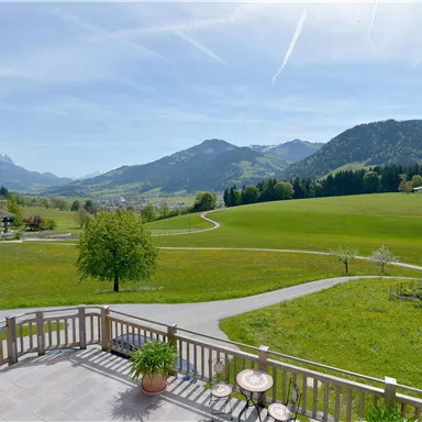 Eine malerische Landschaft mit grünen Wiesen und sanften Hügeln. Im Hintergrund sind Berge und ein klarer, blauer Himmel zu sehen.