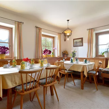 Ein gemütlicher Essbereich mit Holzmöbeln und Tischdecken. Große Fenster bieten einen Blick auf die Berge und frische Blumen dekorieren das Zimmer.