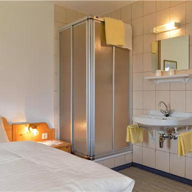 Ein einfaches Zimmer mit einem Bett, einem Bad mit Duschkabine und einem Waschbecken. Helle Wände und freundliche Farben sorgen für eine einladende Atmosphäre.