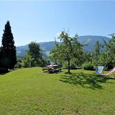 A green meadow with trees and a beautiful view of the mountains. Two deck chairs and a picnic table invite relaxation.