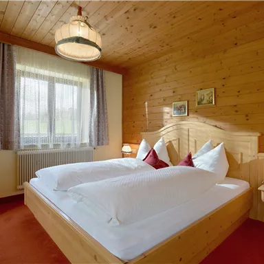 Ein gemütliches Schlafzimmer mit einem Holzbett und weißen Kissen. Die Wände sind aus Holz und es gibt große Fenster, die viel Tageslicht hereinlassen.