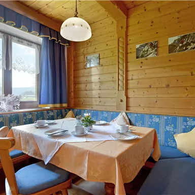 Ein gemütlicher Essbereich in einer Holzstube mit einem Tisch und blauen Sitzkissen. Die Wände sind aus Holz, und das Fenster lässt viel Tageslicht herein.