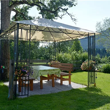 Ein schattiger Pavillon mit Holzmöbeln steht auf einer grünen Wiese. Im Hintergrund sind Bäume und sanfte Hügel zu sehen.