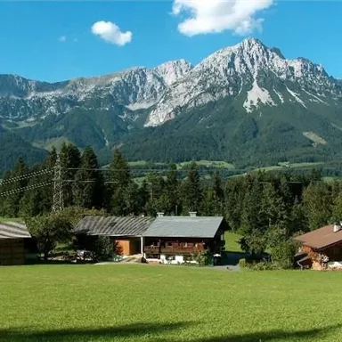 Eine malerische Landschaft mit grünen Wiesen und traditionellen Hütten. Im Hintergrund erheben sich majestätische Berge unter einem klaren blauen Himmel.