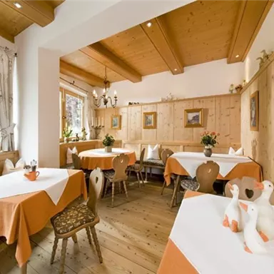 A cozy restaurant with wood paneling and tables covered in orange tablecloths. There are decorative elements such as goose figures and flower arrangements.