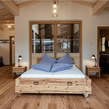Ein stilvolles Schlafzimmer mit einem Holzbett und zwei blauen Kissen. Der Raum hat große Fenster und warmen Holzboden.