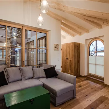 Ein gemütliches Wohnzimmer mit einer grauen Sofaecke und einem grünen Couchtisch. Große Fenster sorgen für viel Tageslicht und einen Blick nach draußen.