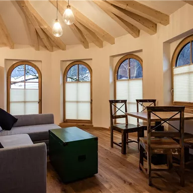 Ein gemütlicher Wohnraum mit einem grauen Sofa und einem grünen Tisch. Große Fenster mit Rundbögen sorgen für viel Tageslicht.