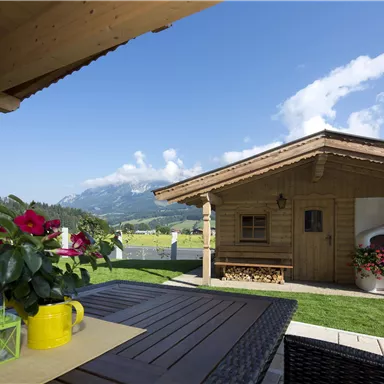 Ein gemütliches Holzhaus in einer malerischen Landschaft. Der Tisch ist mit Blumen und bunten Töpfen dekoriert, und der Himmel ist klar und blau.