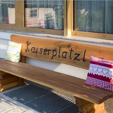 Eine Holzbank mit der Aufschrift "Kaiserplatzl" steht vor einem Fenster. Auf der Bank liegen zwei dekorative Kissen.