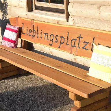 Eine Holzbank mit der Aufschrift "Lieblingsplatz" und bunten Kissen. Der Hintergrund zeigt eine Holzfassade und Pflanzen.