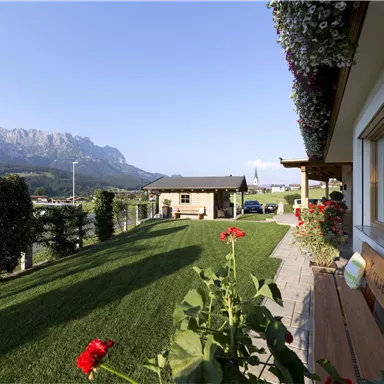 Eine malerische Landschaft mit grünen Wiesen und Bergen im Hintergrund. Bunte Blumen und ein gepflegter Garten runden das Bild ab.