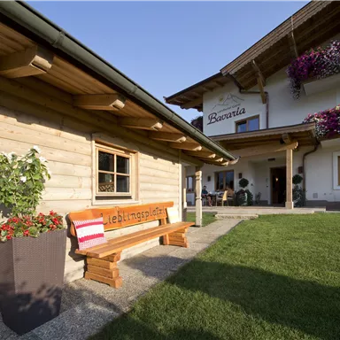 Ein gemütliches Holzhaus mit einer Bank im Vordergrund. Blumen schmücken die Fassade des Hauses und der Rasen ist gepflegt.