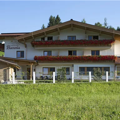 Ein charmantes Gästehaus mit blühenden Balkonkästen und einem gemütlichen Holzaneignung. Umgeben von einer grünen Wiese und Bergen im Hintergrund.