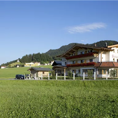 Ein charmantes Haus in ländlicher Umgebung mit grünen Wiesen und Bergen im Hintergrund. Der Himmel ist klar und blau.