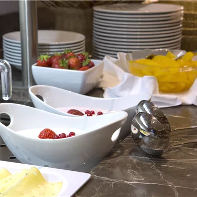 Ein Frühstückstisch mit frischen Früchten, Joghurt und Milch. Im Hintergrund sind weitere Geschirrteile und Obstschalen zu sehen.