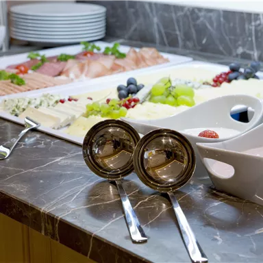 Ein schön angerichteter Buffet-Tisch mit verschiedenen Speisen wie Fisch, Obst und Käse. Im Vordergrund sind zwei glänzende Schalen und Löffel zu sehen.