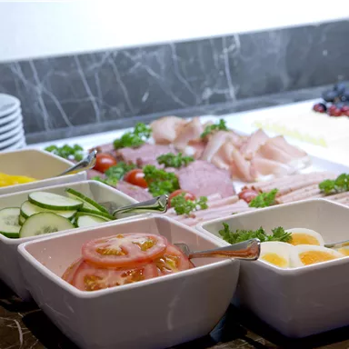 Ein Buffet mit frischen Zutaten, darunter Wurst, Käse, und verschiedene Gemüsesorten. Farbige Schalen mit Gurken, Tomaten und Eiern ergänzen das Angebot.