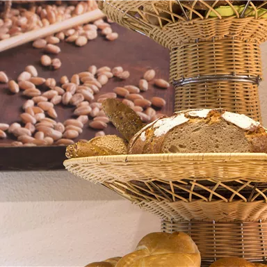 Ein Korb mit frischem Brot und Brötchen steht auf einem Tisch. Im Hintergrund ist ein Gemälde mit Getreidekörnern zu sehen.