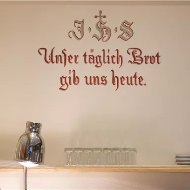 Eine Wand mit der Inschrift "Unser täglich Brot gib uns heute" in dekorativer Schrift. Daneben stehen ein Krug und einige Tassen.