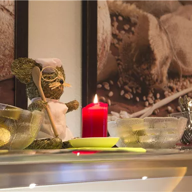 Eine dekorative Küchenzeile mit einer kleinen Figur eines Kochs, einer roten Kerze und mehreren durchsichtigen Schalen. Im Hintergrund sind Bilder von Brot und Getreide zu sehen.