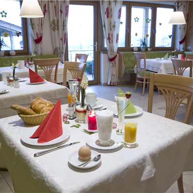 Ein gemütlicher Frühstücksraum mit gedeckten Tischen. Die Tische sind dekoriert mit roten und grünen Servietten, dazu gibt es frisches Brot und Getränke.