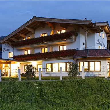 Ein gemütliches Haus mit Holzverkleidung, beleuchtet bei Dämmerung. Umgeben von grünen Wiesen und Bergen im Hintergrund.