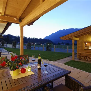 Eine gemütliche Terrasse mit einem Holztisch und Weingläsern. Im Hintergrund sind die Berge und ein Holzhaus bei Dämmerung zu sehen.