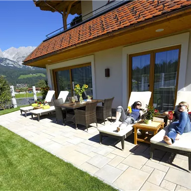 Eine ruhige Terrasse mit Liegen und einem Esstisch im Freien. Im Hintergrund sind grüne Wiesen und Berge zu sehen.