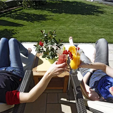 Zwei Frauen entspannen auf Liegestühlen im Freien und stoßen mit bunten Cocktails an. Der Rasen ist grün und die Atmosphäre ist sonnig und entspannt.