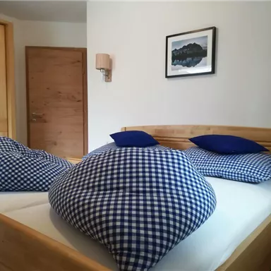 A cozy bedroom with a double bed and large checked cushions. The walls are light and there is a wooden wardrobe as well as a picture on the wall.