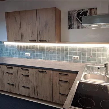 A modern kitchen with wood cabinets and a gray countertop. The walls are equipped with blue tiles and there is a cooktop as well as a sink.