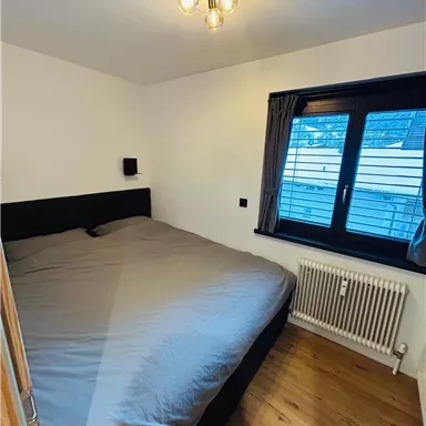 Ein einfaches Schlafzimmer mit einem großen Bett und einem Fenster. Die Wände sind hell und der Boden ist aus Holz.