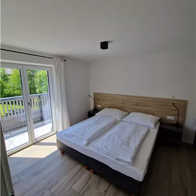 Ein modernes Schlafzimmer mit einem Doppelbett und großen Fenstern. Der Raum ist hell und freundlich mit Holzfußboden und Zugang zu einem Balkon.