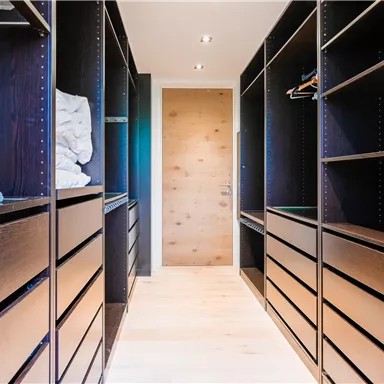 Ein leerer Ankleideraum mit offenen Regalen und einer Holztür. Der Boden ist hell und der Raum wirkt modern und geräumig.