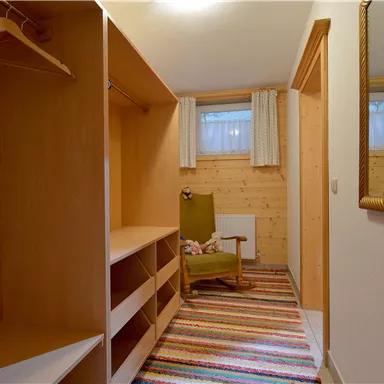 Ein gemütlicher Flur mit einem Holzregal und einem bequemen Sessel. Der Boden ist mit einem bunten Teppich ausgelegt, und ein Spiegel hängt an der Wand.