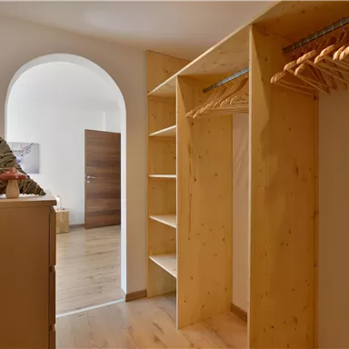 Ein heller Eingangsbereich mit einem offenen Kleiderschrank aus Holz. Im Hintergrund ist eine Tür sichtbar, die zu einem weiteren Raum führt.