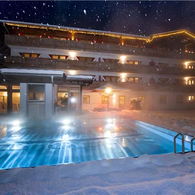 Ein gemütliches Hotelgebäude im Schnee mit einem beleuchteten Pool. Der Nachthimmel ist klar und sternenklar.