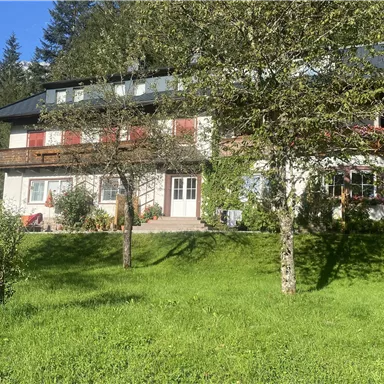 Ein schönes Haus mit Balkon, umgeben von grünem Rasen und Bäumen. Im Hintergrund sind Berge zu sehen.