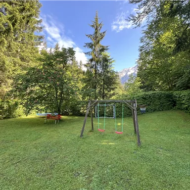 Ein grüner Garten mit einer Schaukel und einem Tisch. Umgeben von hohen Bäumen und einem blauen Himmel.